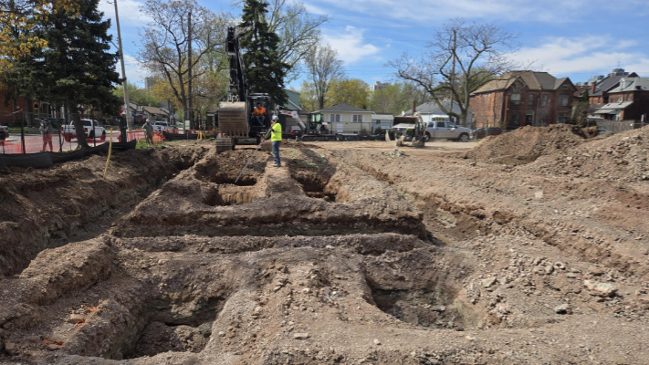 A construction site with deep trenches, machinery, and workers in safety gear, surrounded by trees and residential buildings.