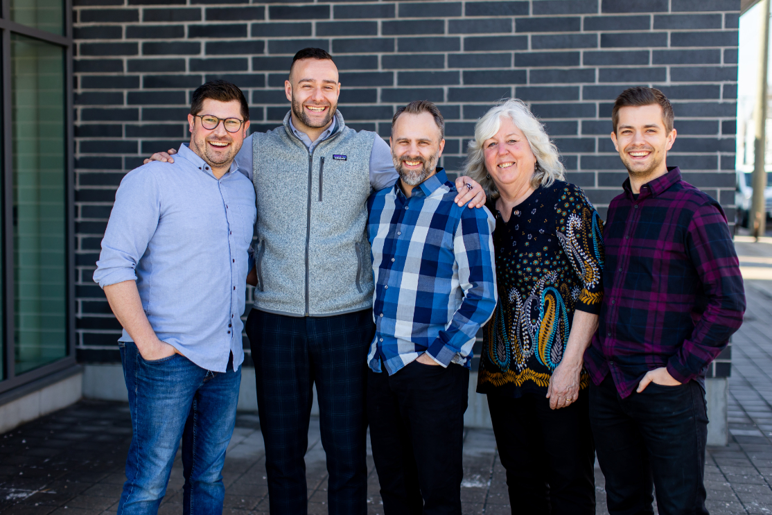 A group of five people stands together outside a building, smiling and dressed in casual attire.