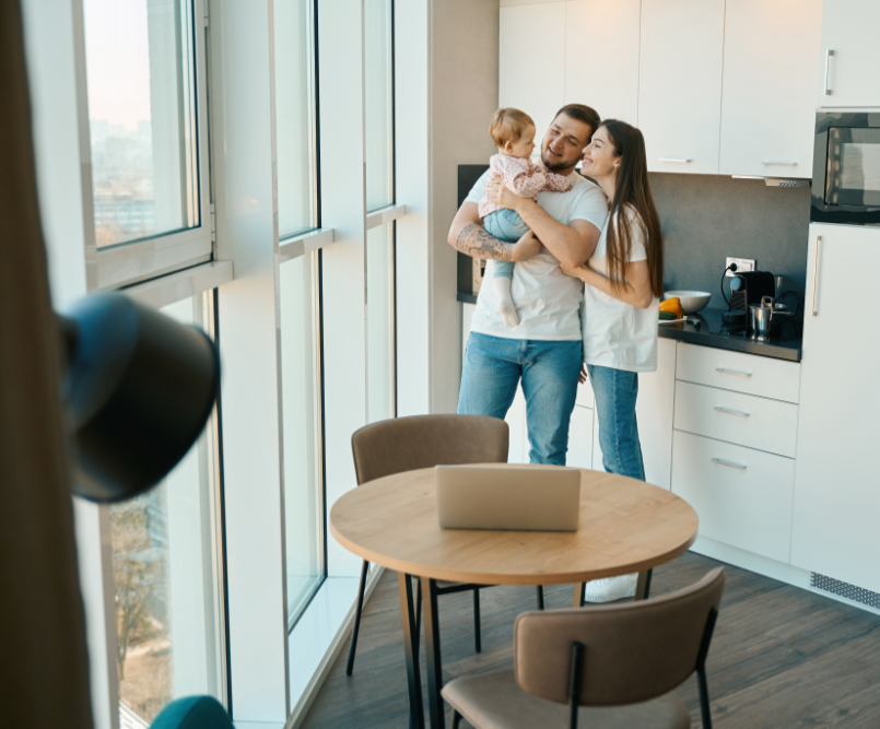 A happy family of three stands in a modern kitchen, with a baby in the father's arms, smiling at each other. A laptop sits on the table.